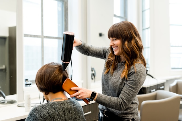 A stylist blow-drying a client's hair in a bright, window-lit salon
